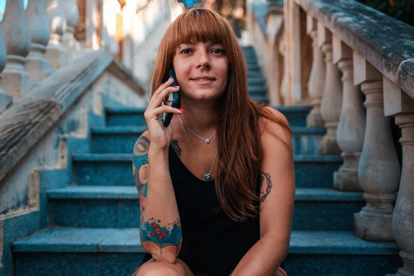 タトゥーのある若い笑顔の女性の肖像画は 石の階段に座って電話で話しています コミュニケーションとビジネスのコンセプト — ストック写真