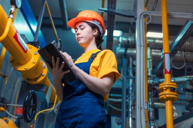 Üniformalı ve koruyucu miğferli genç bir kadın işçinin portresi elinde dijital tabletle ekipmanları denetliyor. Alt manzara. Endüstri kavramı.