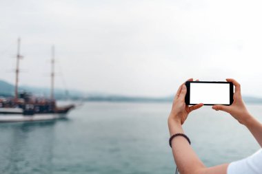 Model yap. Kadın telefonu yatay olarak tutuyor. Arka planda, deniz ve gemiler. İzole et.