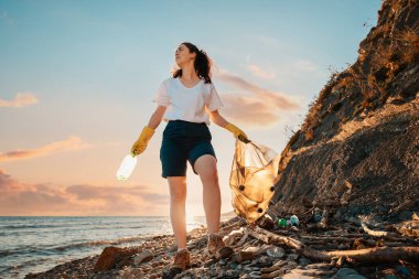 Kadın bir aktivist elinde plastik bir torba ve elinde bir şişeyle gün batımının arka planında duruyor. Alt manzara. Kıyı şeridini ve vahşi plajları temizliyorum. Dünya Günü konsepti.