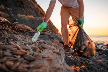 Dünya çevresi günü. Erkek bir aktivist plajdan plastik bir şişe alıyor. Arka planda, deniz ve günbatımı. Düşük açı. Sahil temizliği ve Dünya Günü kavramı..