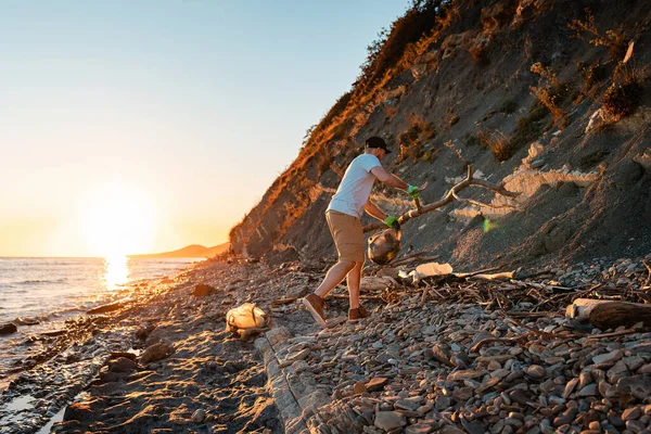 World environment day. A male activist is cleaning up on a wild beach. In the background, the sea and the sunset. The concept of cleaning the coastal zone.