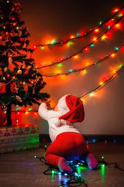 Christmas. A cute baby in a Christmas hat crawls around a Christmas tree decorated with glowing lights. Rear view. The concept of winter Christmas holidays.