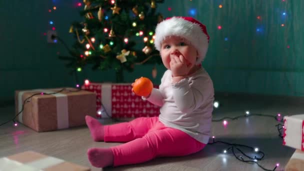 戴着圣诞礼帽的小女孩坐在圣诞树旁吃橘子 慢动作新年假期的概念 — 图库视频影像