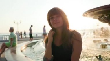 Kolunda dövmesi olan güzel, bronzlaşmış genç bir kadın çeşmenin yanında oturuyor. Arka planda gün batımı. Gerçek zamanlı. Yaz tatili kavramı.