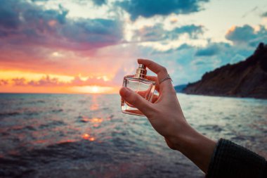 Kadın eli, altın şeffaf parfüm şişesini spreyliyor. Yakın plan. Arka planda, okyanus ve bulutlu gün batımı. Parfüm reklamı kavramı.
