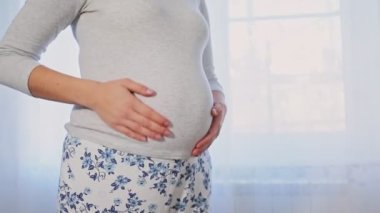 Genç hamile bir kadın pencerenin önünde durur ve karnını okşar. Elleriyle kalp yapıyor. Karnı ve elleri yakın çekim. Hamilelik ve annelik kavramı.