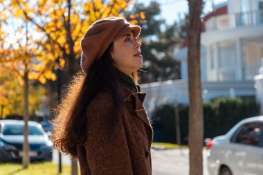 Güneşli bir şehirde sonbahar caddesinde yürürken şapkası ve paltosu olan güzel, genç bir kadının yan görüntüsü. Psikoloji kavramı ve pozitif ruh hali.