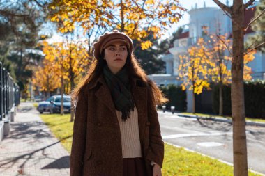 Güneşli bir şehirde, sonbahar caddesinde yürüyen, eski moda şapka ve palto giyen üzgün, genç bir beyaz kadının orta fotoğrafı. Psikoloji kavramı.