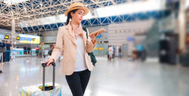 Zarif takım elbiseli beyaz bir kadının geniş açılı resmi akıllı telefon kullanıyor ve bavul tutuyor. Havaalanında biri var. Uzayı kopyala Web seyahat ve seyahat bayrağı.