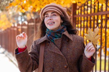 Dişleri askılı, ceketli ve şapkalı genç, güzel beyaz kadın portresi. Sonbahar sarısı yaprakları ve sokakta el sallayan el sallıyor. Yaşam tarzı ve şehir hayatı