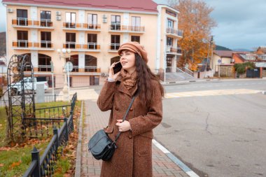Beyaz kadının ceketli ve şapkalı gülümseyen bir şekilde akıllı telefondan aradığı bir fotoğraf. Arka planda sonbahar şehri.