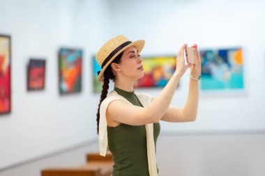Zarif takım elbiseli beyaz bir kadının yan görüntüsü bir sanat galerisinde ve internet gezisi için akıllı telefonunu kullanıyor. Öğrenciler için müzede modern kültür eğitimi.