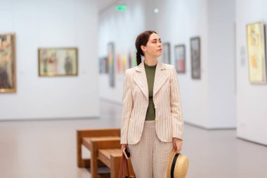 Hasır şapkalı, zarif ceketli ve sanat galerisinde poz veren beyaz bir kadının orta pozu. Arka planda boyama. Kültür Eğitimi ve Müze Günü kavramı.