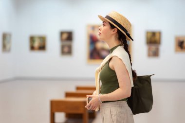 Hasır şapkalı, zarif elbiseli ve galeride modern sanatı düşünen beyaz bir kadının yan görüntüsü. Uzayı kopyala Müze Günü konsepti.