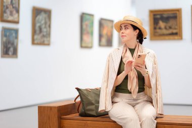 Beyaz tenli güzel bir kadının zarif bir takım elbiseyle bir sanat galerisindeki bankta otururken ve akıllı telefonunu kullanırken görüntüsü. Uzayı kopyala Modern kültür eğitimi.