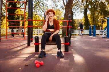 Orta yaşlı, mutlu, beyaz, kıvırcık saçlı kadın spor salonunda oturuyor ve cep telefonuyla arıyor. İletişim kavramı.