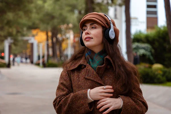 Bereli ve ceketli güzel bir kadın, bir sonbahar günü şehir sokaklarında dolaşırken kulaklıklarıyla müziğin keyfini çıkarıyor ve dinlenmek için zaman ayırıyor.