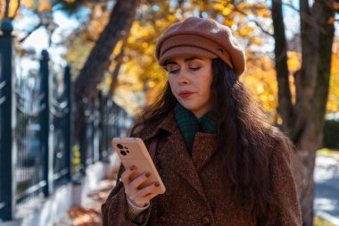 Zarif, üzgün, düşünceli, beyaz bir kadının ceketli ve şapkalı akıllı telefon kullanışının orta fotoğrafı. Arka planda güneşli güzel bir cadde var. Modern iletişim ve sosyal medya.