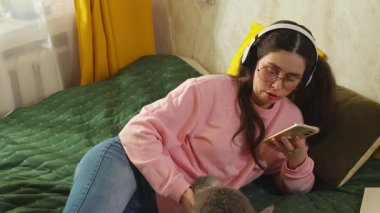 Genç, beyaz bir kadın yatağında oturmuş, tavşanını okşuyor ve sessiz bir ev gecesinin tadını çıkarıyor. Akıllı telefon kullanan bir kız. Hayvanlar, hayvan bakımı ve yaşam tarzı kavramı