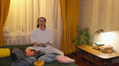 Kafkas görünümlü genç bir kadın valizini tatil için hazırlarken yatağında kişisel eşyalarını düzenliyor. Seyahat paketi ve ev organizasyonu kavramı. Geniş açı, boşluğu kopyala