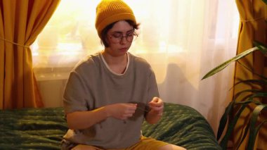 Genç, beyaz bir kadın renkli ipliklerle iğne ipliğinde yatağında oturmuş nakış işine hazırlanıyor ve rahat yatak odasının içinde sakin yaratıcı bir hobiyle eğleniyor. Orta çekim.