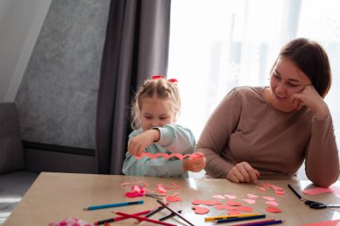 Genç, mutlu beyaz bir anne ve küçük tatlı kızı kırmızı kağıttan kalp kesiyorlar. Anneler Günü ve Sevgililer Günü konsepti.