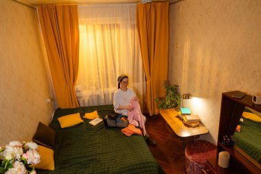 Kafkasyalı genç bir kadın gün batımında iç mekânda kıyafet düzenliyor. O, giysileri sakince katlar. Düzenli ev rutini. Geniş açı.