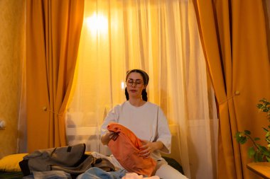 Genç beyaz kadın gün batımına kadar yatak odasında giysilerini katlıyor. O, düzenli bir oda tutar. Evde bakım konsepti. Düşük açı görünümü,