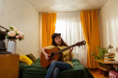 Beyaz, genç bir kız öğrenci evinde yatağında otururken gitar çalıyor. Ev müziği öğrenme süreci. Yaratıcı hobi ve sanatsal beceri geliştirme. Alt görünüm.