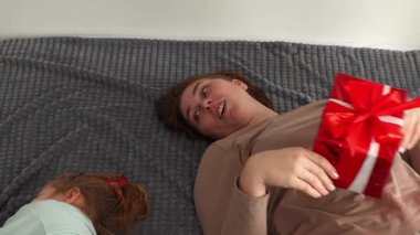 Kafkas anne ve kızının üst görünüşü bayram için bir hediye tutuyor ve yatakta uzanıyor. Anaokulu eğitimi ve Sevgililer Günü konsepti.