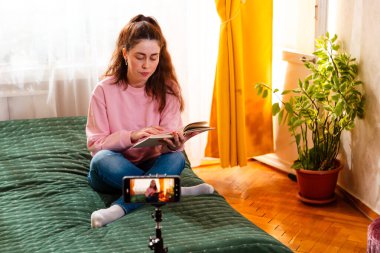 Genç bir kadın yatakta oturup dijital takipçileri için bir video bloğu kaydederken kendini akıllı telefondan çekiyor. Orta çekim.