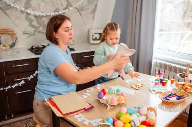 Mutfak masasında annesiyle otururken küçük bir kızın tavşan eldiveniyle oynamasını geniş açıdan görmek. Çocuk Paskalya oyunları oynuyor. Paskalya Bahar Festivali.