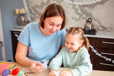 Genç ve güzel bir annenin portresi ve küçük tatlı bir kız Paskalya bahar tatili için el sanatları süsler. Aile mutfaktaki masada oturuyor..