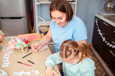 Genç ve güzel bir anne ve küçük tatlı bir kızın Paskalya bahar tatili için el sanatları çalışmalarının en güzel manzarası. Aile mutfaktaki masada oturuyor..