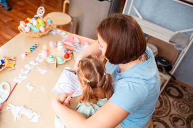 Genç bir anne, kızının masada kağıt Paskalya süslerini kesmesine yardım eder. Evde yapılacak aile tatiline hazırlanırken birlikte kaliteli zaman geçirir. Üst görünüm.