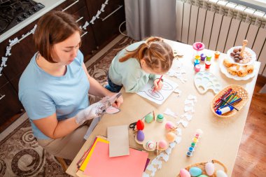 Gülümseyen bir anne ve kız evde oturup mutlu bir bahar aile geleneğinin parçası olarak el yapımı Paskalya süslemeleri yapıyorlar. Üst görünüm.