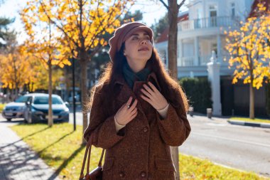Güneşli bir şehirde sonbahar caddesi boyunca şapkası ve paltosuyla yürüyen güzel bir genç kadının orta fotoğrafı. Mutluluk ve pozitif ruh hali kavramı.