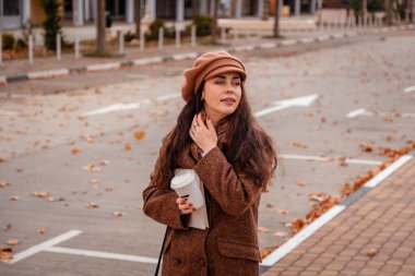 Sonbahar renklerinde bir kadın sessiz bir şehir yolunda sıcak kahvenin tadını çıkarıyor. Yumuşak rüzgar, ılık içecek ve hafta sonu sadeliği.
