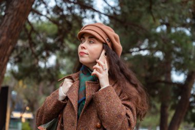 Şapka ve paltolu güzel beyaz bir kadının portresi sokakta yürüyor ve telefonda konuşuyor. Modern iletişim kavramı.