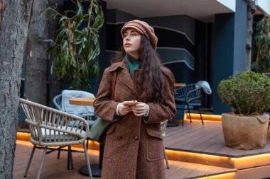 Paltolu ve bereli güzel beyaz kadın akıllı telefonuyla mesajlaşırken ve huzurlu bir hafta sonunun tadını çıkarırken sonbahar şehri sokaklarında yürüyor..