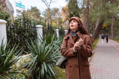 Şık paltolu ve bereli güzel beyaz kadın şehir sokaklarında yürüyor, huzurlu şehir manzarasının tadını çıkarıyor. Şehir eğlencesi ve modern yaşam tarzı kavramı