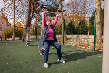 Yeşil bir parkta açık hava fitness makineleri kullanan yaşlı beyaz bir kadın. Fiziksel aktivite, sağlık ve emeklilik hayatı