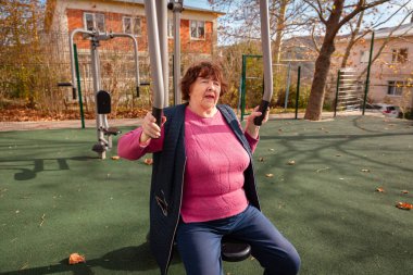 Parktaki açık hava fitness ekipmanları üzerinde yapılan ameliyatlardan sonra iyileşme sürecinde olan son sınıf beyaz kadın. Fiziksel aktivite rehabilitasyonu, gücü ve sağlıklı yaşlanmayı destekler.