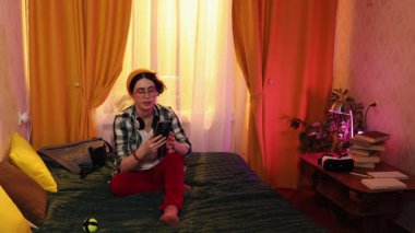 Gündelik giyinen beyaz bayan öğrenci rahat bir şekilde yatakta oturuyor, cep telefonuyla konuşuyor ve evde boş vakitlerin, iletişimin, günlük yaşam tarzının ve gençlik konforunun tadını çıkarıyor. Boşluğu kopyala.