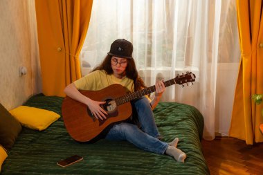 Beyaz, genç bir kız öğrenci evdeki bir yatakta gitar öğreniyor. Kapalı alanda müzik alıştırması. Yaratıcılık ve eğitim. Üst görünüm.