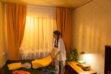 Beyaz bir kadın gün batımında yatak odasında çamaşır katlıyor. O, düzenli depolamaya odaklanıyor. Ev rutini konsepti. Üst manzara, geniş açı.