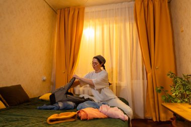 Gün batımının sakin olduğu saatlerde genç beyaz kadın içeride kıyafet katlıyor. Yaşadığı yeri düzenliyor. Ev temizliği konsepti. Düşük açı, geniş açı..