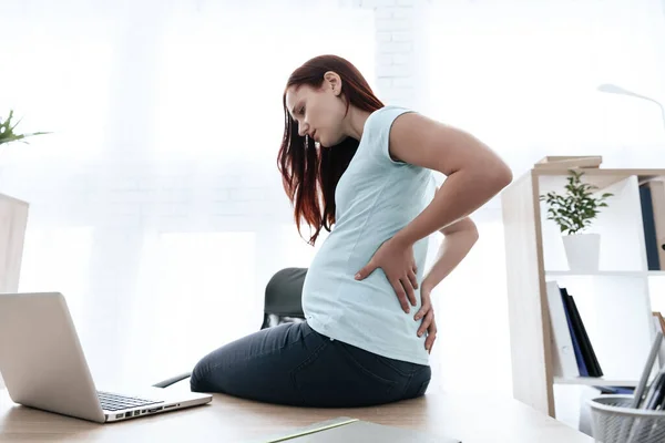 A woman sits on a table and grabs her sore back.