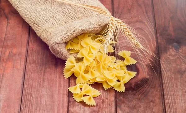 Uncooked bow-tie pasta and wheat spikes on wooden surface